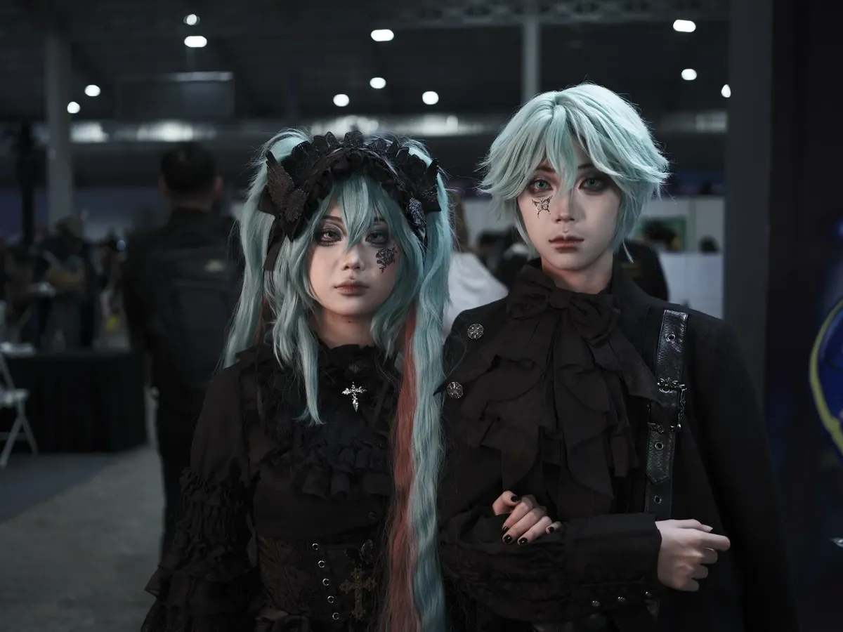 Two individuals in gothic cosplay at an indoor anime convention. Unique and vibrant costumes.